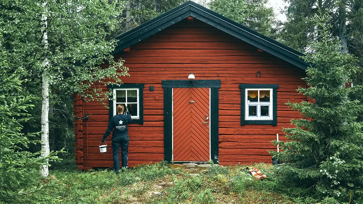 En person som håller en hink står framför ett rött hus omgivet av gräs och träd.
